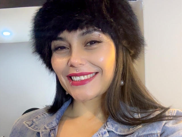 CharlotteCrofort con capelli castani, cappello nero di pelliccia, rossetto rosso, piercing al naso, che sorride, giacca di jeans, orecchini di perle, glutei visibili, carina, attraente, pelle chiara, leggero rossore, espressione felice..