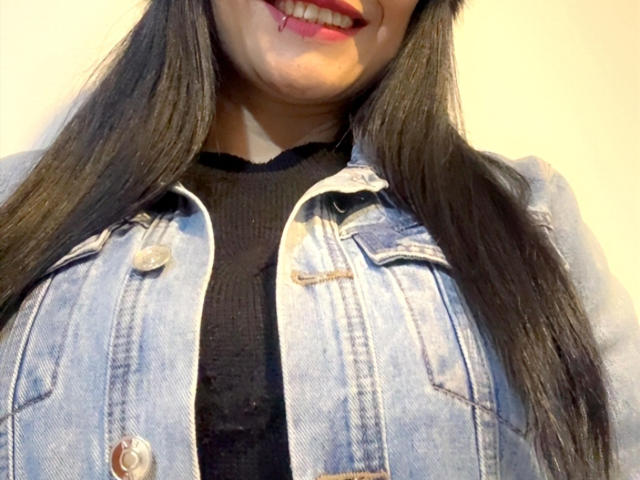 CharlotteCrofort with long black hair, smiling with red lipstick, wearing a light blue denim jacket over a black rib-knit top, showing her big boobs.