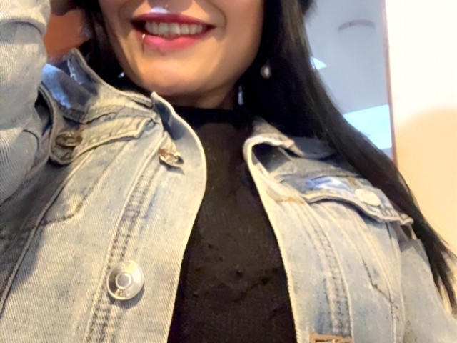 CharlotteCrofort's face, wearing a denim jacket, black sheer top, and pink lipstick, with long black hair and small pearl earrings, smiling slightly, showing a small boop on her nose.