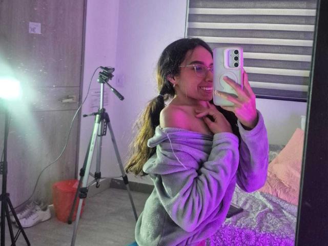MiliBlom with long brown hair in a ponytail, glasses, and a gray off-shoulder hoodie, taking a mirror selfie, showing her bare shoulder and smiling. TriPod and light in background.