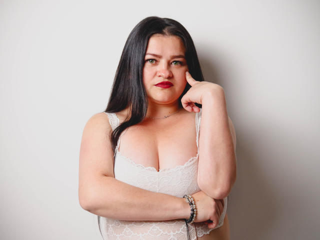 CamilaGiamcomo, con lunghi capelli neri, rossetto rosso, top bianco in pizzo, che si tocca la guancia, mostrando il seno, braccialetto al polso destro, che guarda la fotocamera, sfondo bianco..