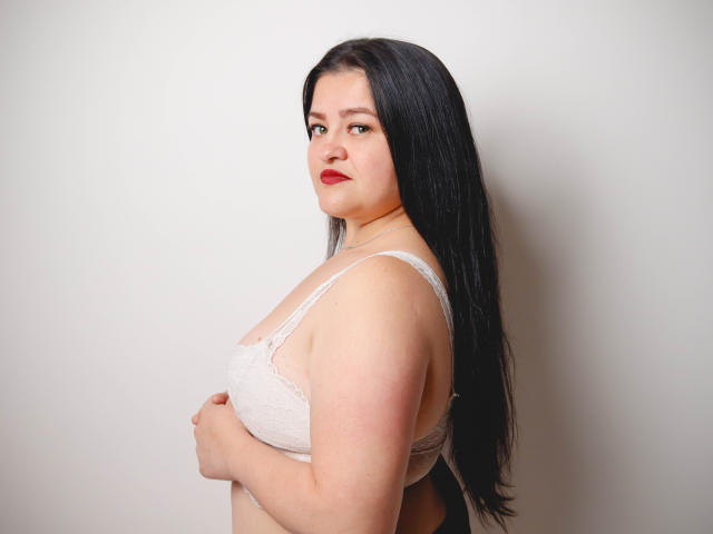 CamilaGiamcomo with long black hair, wearing a white lace bra, holding her tits, looking over her shoulder with red lipstick, standing against a plain wall.