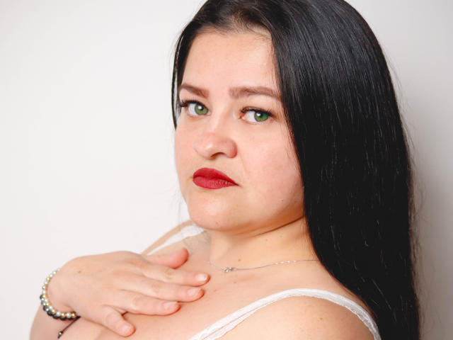 CamilaGiamcomo with black hair, green eyes, red lipstick, white top, hand on chest, bracelet, and necklace, looking slightly to the side, showing a confident, slightly sassy expression.