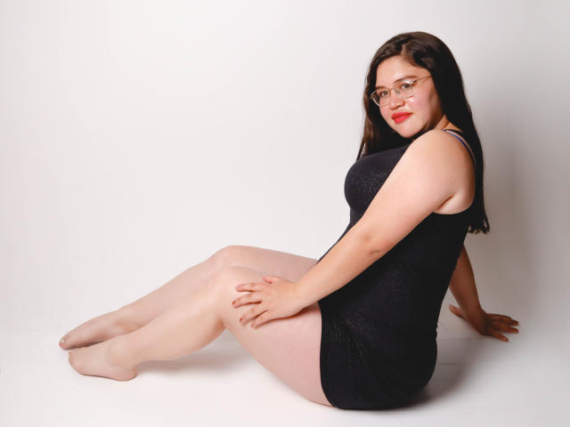 AliceGrylls with glasses, black sparkly dress, and pale skin, sitting with legs out, hands on thighs, red lipstick, black hair, curvy with a round ass, looking at camera.