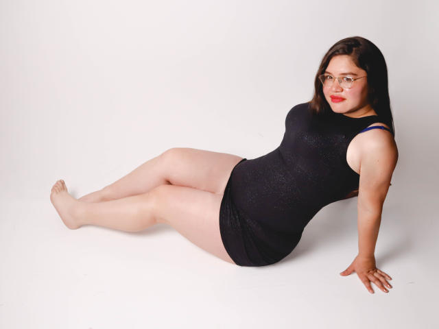 AliceGrylls, a chubby woman with pale skin, red lipstick, and glasses, is sitting on the floor, legs stretched out, wearing a black, sparkly, sleeveless dress, showing her ass.