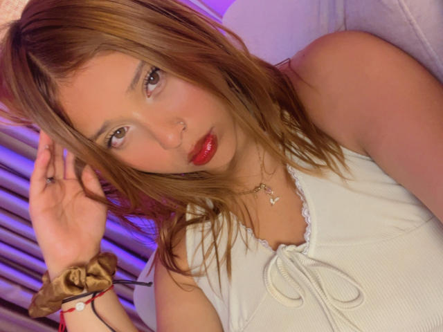 HeraJuno with wet brown hair, red lips, white sleeveless top, nose ring, and brown scrunchie on wrist, looking at camera with hand touching hair. Her tits are small.