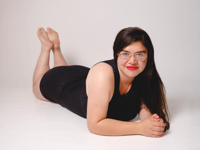 AliceGrylls is lying on her stomach, wearing a black sleeveless dress, with her ass up, feet in the air, and glasses on, smiling with red lipstick.