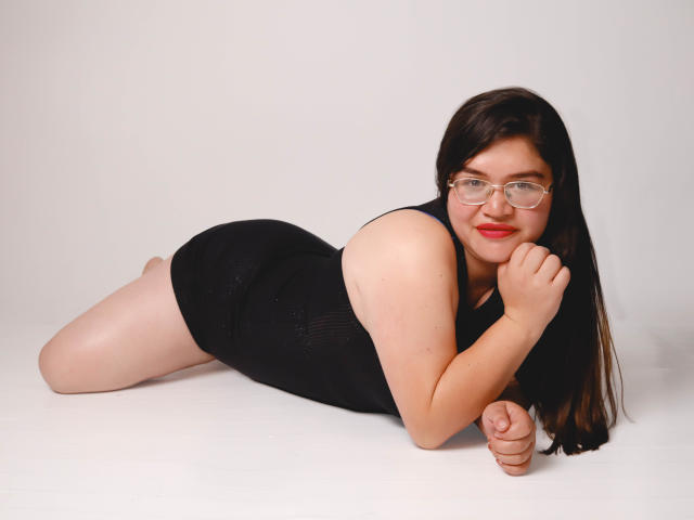AliceGrylls, lying on her stomach, wearing a black tank top and shorts, with glasses, smiling, showing her ass, and her hand near her mouth.