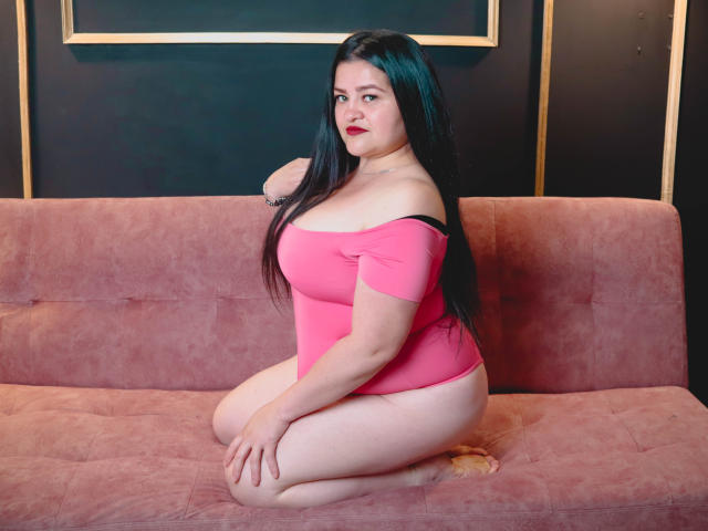 CamilaGiamcomo a de grandes fesses, une peau pâle, des cheveux noirs, du rouge à lèvres rouge, et elle porte un haut rose à une épaule, étant agenouillée sur un canapé en suède rose..
