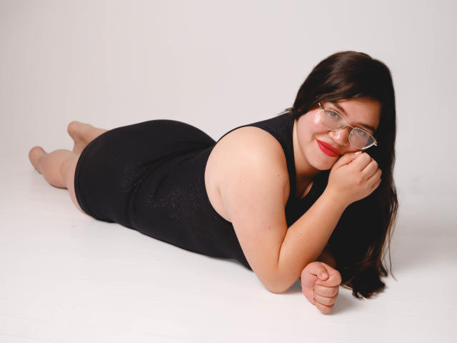 AliceGrylls is lying on her stomach, wearing a black dress, glasses, and red lipstick, with her ass up and her tits slightly visible, smiling at the camera.