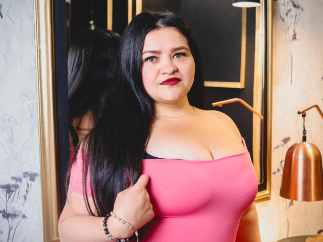 CamilaGiamcomo with long black hair, pink off-shoulder top showing her big boobs, red lipstick, and bracelet, standing in front of a mirror, looking at the camera.
