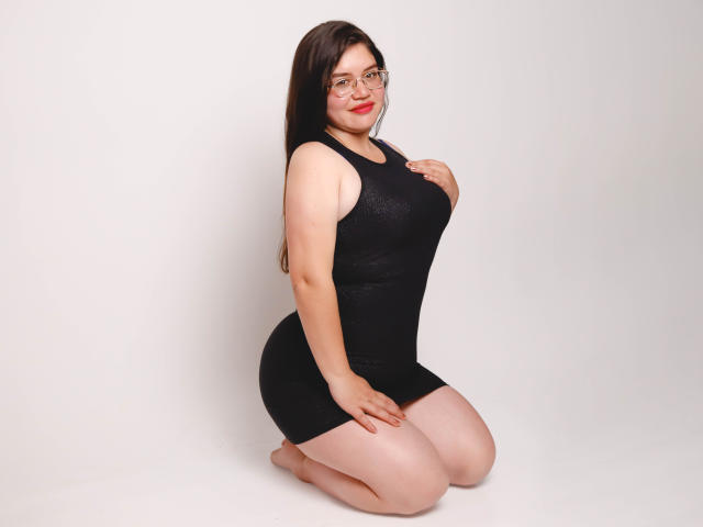 AliceGrylls, kneeling, wearing a tight black dress, glasses, and red lipstick, with her hand on her chest, showcasing her big ass and curvy body.