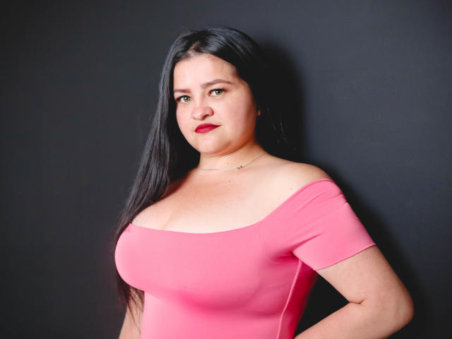 CamilaGiamcomo with large tits, wearing a tight pink off-shoulder top, black hair, red lipstick, and a subtle necklace, standing against a dark background.
