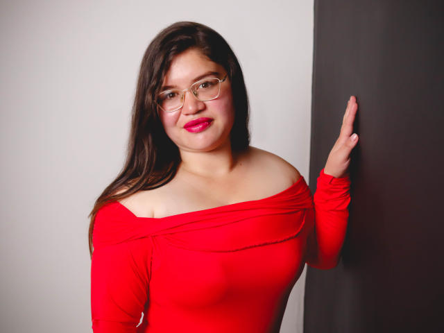 AliceGrylls with long dark brown hair, glasses, red off-shoulder top, and pink lipstick, standing against a wall, hand raised, showing her bare shoulders and small tits.