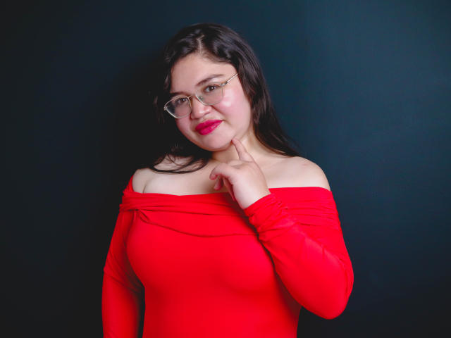 AliceGrylls with red off-shoulder top, glasses, and pink lipstick, touching chin with right hand, wearing red dress, dark hair, and red lipstick, against dark background.