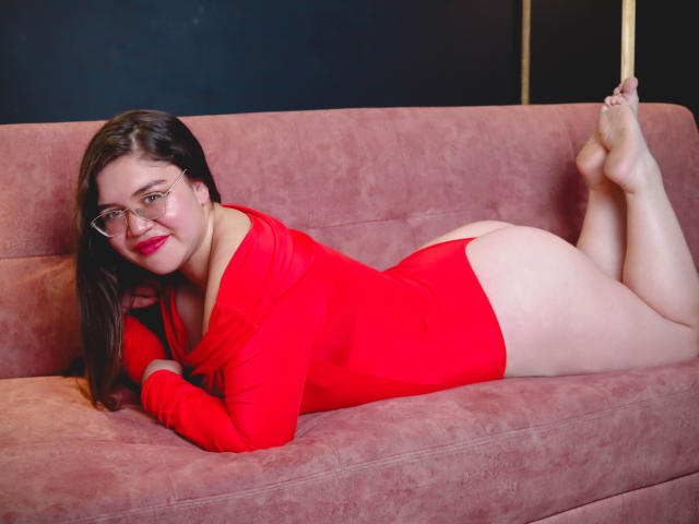AliceGrylls lying on a pink couch, wearing a red one-piece, her ass up, feet in the air, glasses, red lipstick, smiling, brown hair, curvy, white skin.