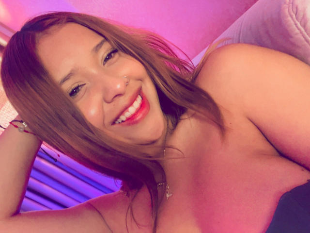 HeraJuno with brown hair, smiling, nose piercing, red lipstick, bare shoulders, necklace, pink and purple lighting, arm raised, smiling teeth, cute, sexy, attractive.