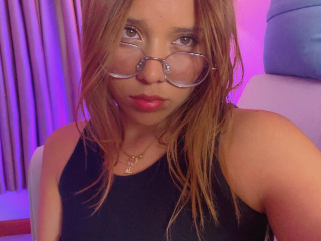 HeraJuno with wet brown hair, glasses, red lipstick, black tank top, showing her bare shoulders and upper chest, looking into the camera with a slight pout.