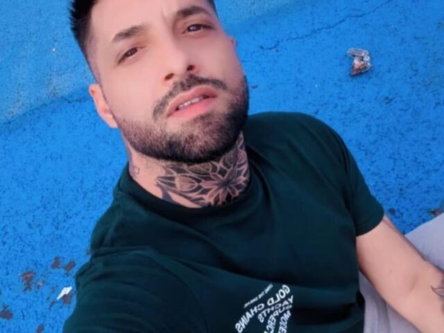 TommyDum, with a beard and tattoo on his neck, wearing a green shirt, has short black hair and a slightly open mouth, sitting on a blue surface.