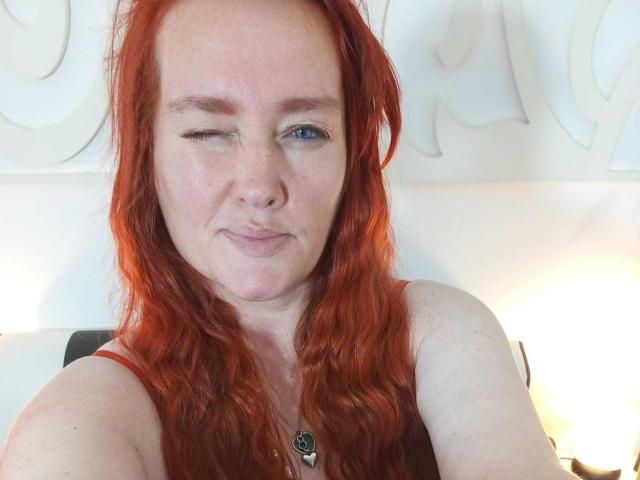 SinFulTitsDD a les cheveux roux ondulés, une peau claire, des yeux bleus et un clin d'œil espiègle. Elle porte un haut rouge avec un collier vert, et la photo en selfie montre ses fesses..