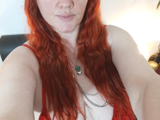 SinFulTitsDD con cabello rojo ondulado, piel clara, vistiendo una blusa roja de encaje, un collar con un dije verde y una cadena de plata, tomándose un selfie, mostrando su pecho y labios fruncidos..