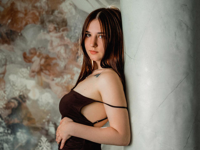MayaWinters with brown hair, sideboob, and small tattoo, leaning against a white column, wearing a brown, strapless dress, looking at the camera with a slight smile.