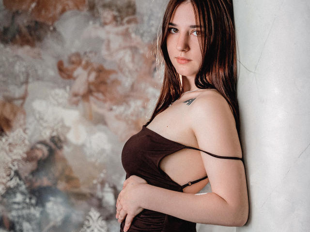 MayaWinters with long brown hair, side-eyeing camera, wearing a brown, cut-out, side-baring top, exposing her small tits and a tiny tattoo on her right shoulder.