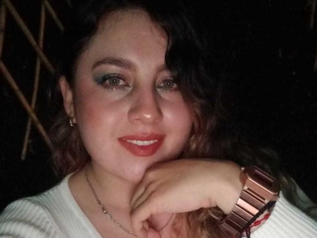 Alicelebell hat lockiges, braunes Haar, grünen Lidschatten, roten Lippenstift, ein weißes Rippstrickshirt, eine roségoldene Uhr und lächelt dabei, wobei ihr Kinn auf ihrer Hand ruht..