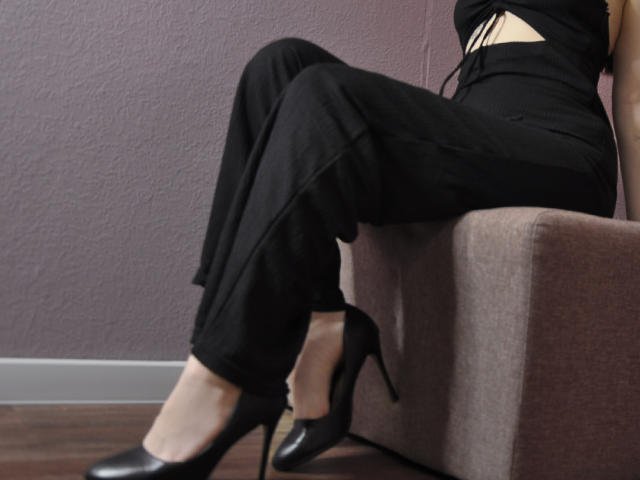 MayaWinters sitting on a chair, wearing black pants, black high heels, and a black top with a cutout, showing a small bit of her ass.