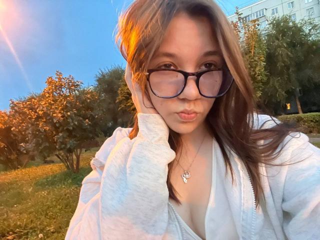 Aurelia Emswiler, avec des lunettes, des lèvres boudeuses, des cheveux bruns, un sweat à capuche blanc, un collier, une main sur la joue, éclairée par la lumière du coucher de soleil, avec un ciel bleu, des arbres et un bâtiment en arrière-plan. Seins visibles..