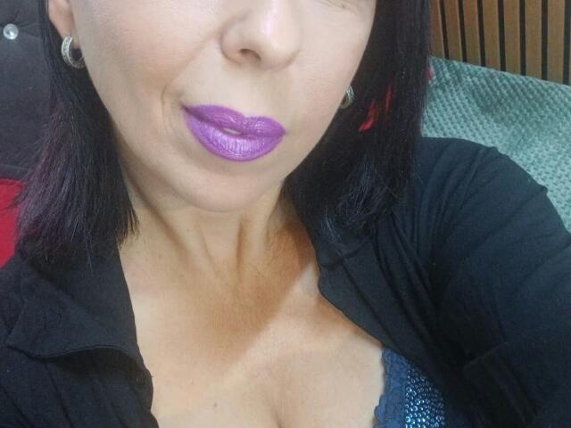 LauraSin with purple lipstick, black shirt, and blue bra showing her tits, dark hair, and earrings, smiling slightly. Her skin is tan, and she's wearing a black cardigan.