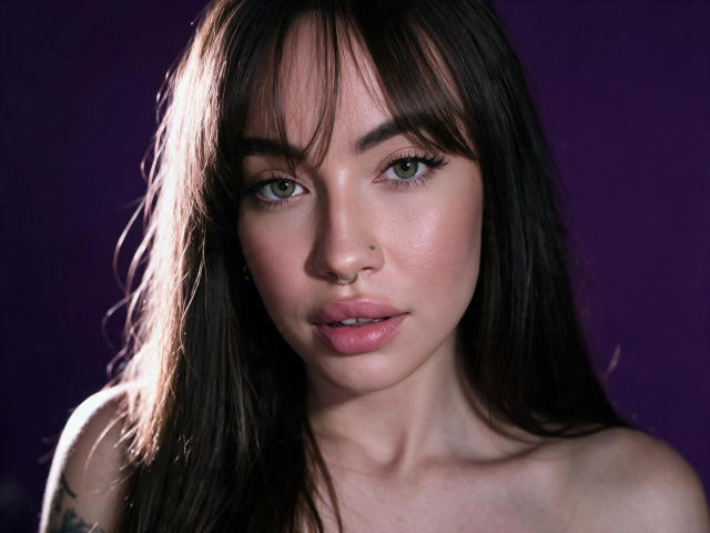 LexiHerzig-hot has long dark brown hair, green eyes, pink glossy lips, a small nose piercing, and a tattoo on her right shoulder. She's looking directly at the camera.