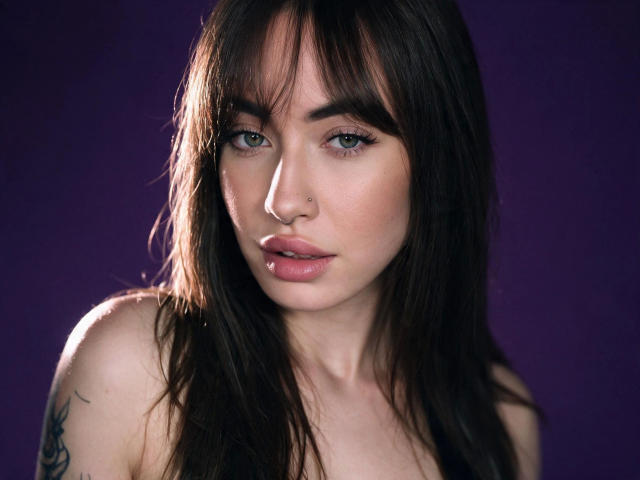 LexiHerzig-hot has dark brown hair, blue eyes, a nose ring, and a lip piercing. She's looking at the camera with a serious expression, showing off her tattooed shoulder.