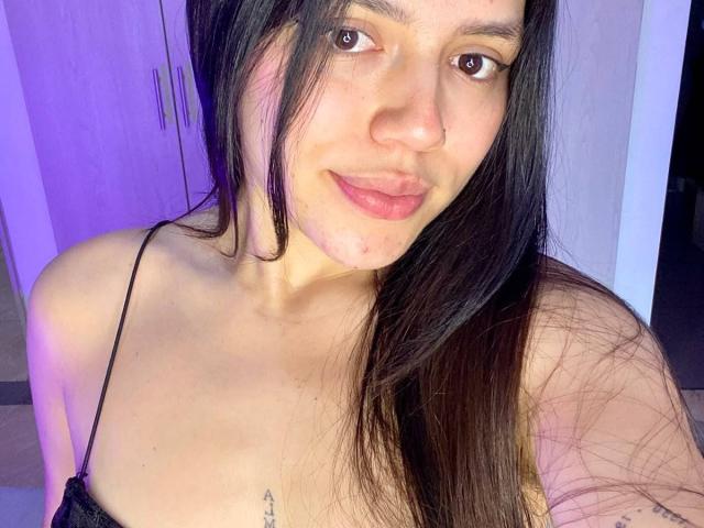 SophiaMarx has long black hair, brown eyes, and a nose ring. She's smiling with pink lips, showing a small tattoo on her chest. She's wearing a black strap top.