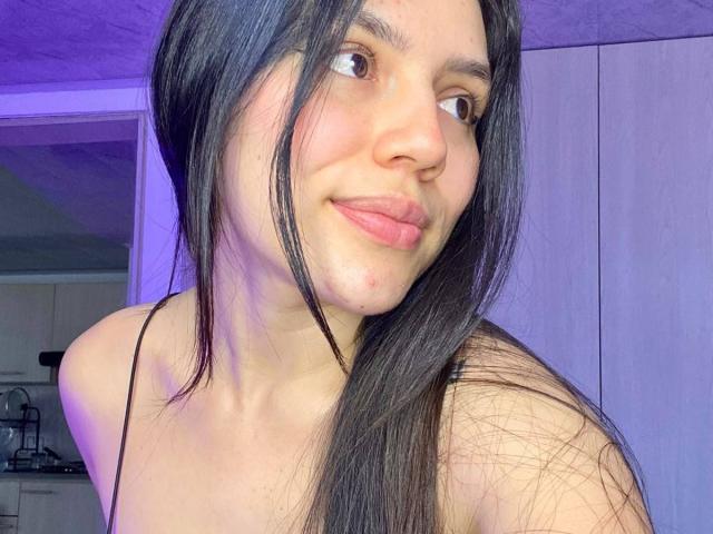 SophiaMarx with long black hair, brown eyes, and pink lips, smiling at the camera, showing her bare shoulders and a small red pimple on her left cheek.
