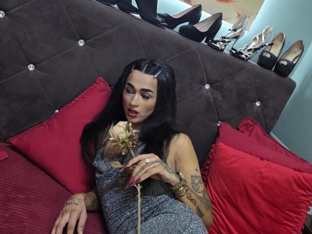 IrisIrs with black hair, red lipstick, tattoos, and a grey dress, holding a rose, laying on a brown, tufted, suede couch with red pillows, and high heels in background.