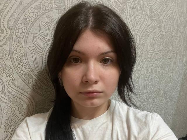 Deedra Brunscheen a la peau pâle, les yeux marron et les cheveux noirs attachés en queue de cheval. Elle porte une chemise blanche, a une expression neutre et regarde directement la caméra..