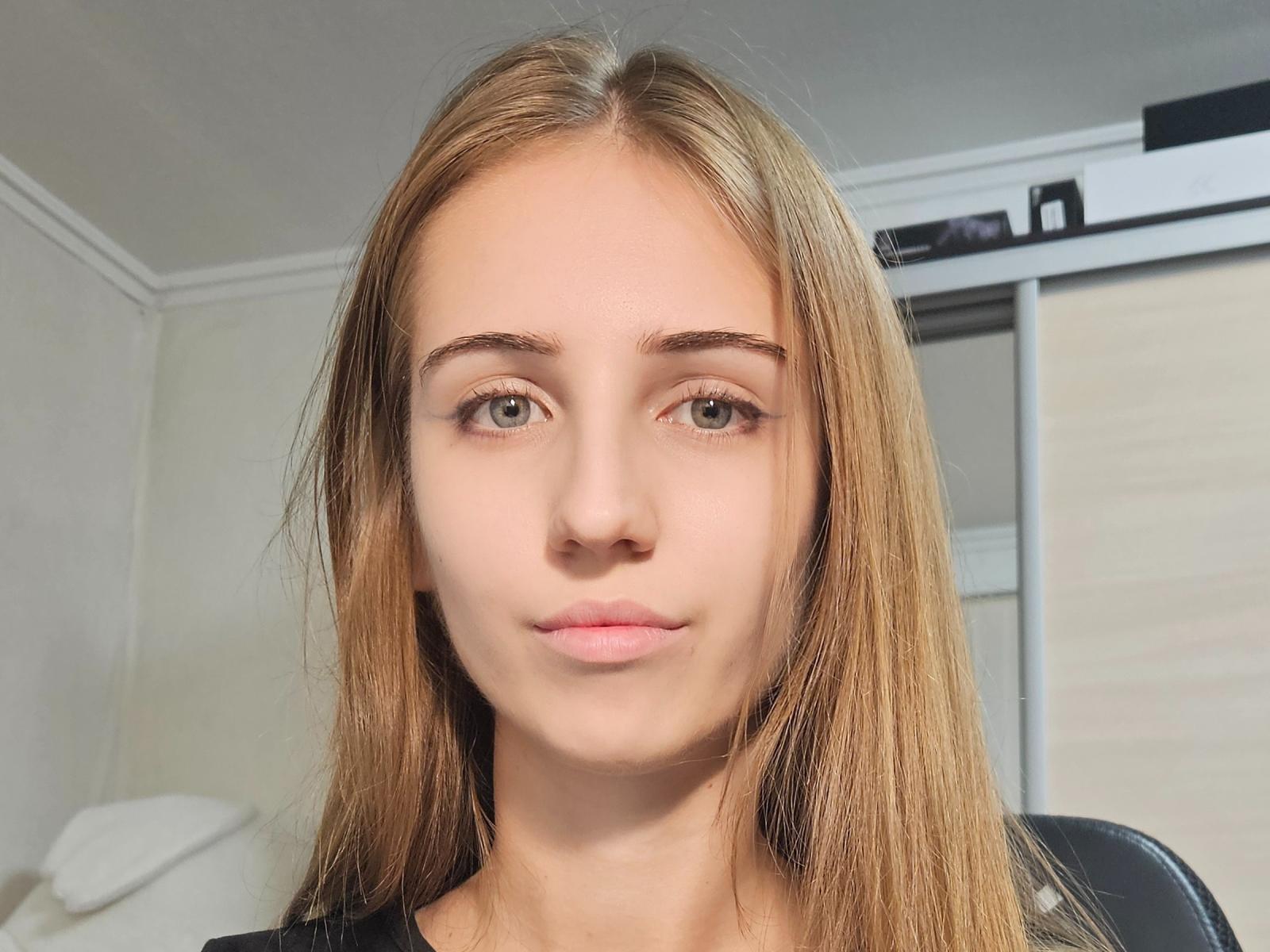 Hilary Escatel tem cabelo castanho claro e liso, olhos verdes e uma expressão neutra. Ela está vestindo uma blusa preta, e uma cadeira preta está levemente visível atrás dela..