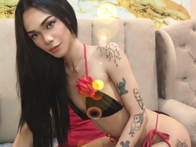 HeavenHunter with long black hair, red and black bikini, tattoos on arms and chest, sitting on beige sofa, looking at camera, soft smile, small lens flare in front.