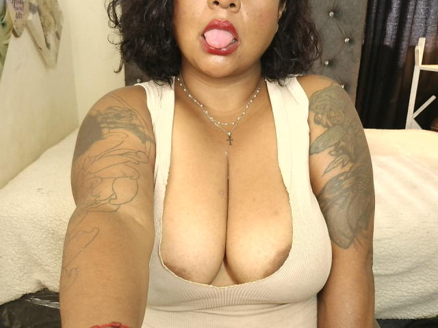 JoanaQueens with tan skin, curly black hair, and red lipstick, sticking out her tongue, wearing a low-cut beige top, showing large tits, and tattoos on her arms.