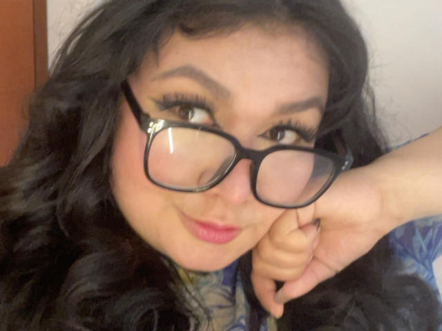 LillyMayer with curly black hair, black glasses, and pink lipstick, resting her chin on her hand, smiling slightly. Her brown eyes are accentuated with bold eyeliner.