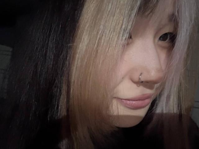 JungMcgranor's face, nose ring, and pale pink lips, with black and blonde hair partially covering her eyes, in a dimly lit photo.