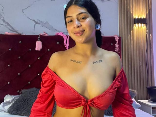 LonxEva with tan skin, black hair, and small tits, wearing a red off-shoulder top, smiling at the camera, has tattoos on her chest, and large ear gauges.