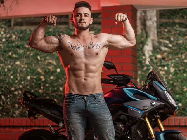 AshtonMuller, musculoso, sem camisa, exibindo os bíceps, com tatuagens no peito, calça jeans azul, em pé em frente a uma motocicleta azul, com um fundo de tijolos vermelhos e trepadeiras verdes, cabelo curto e escuro, barba..
