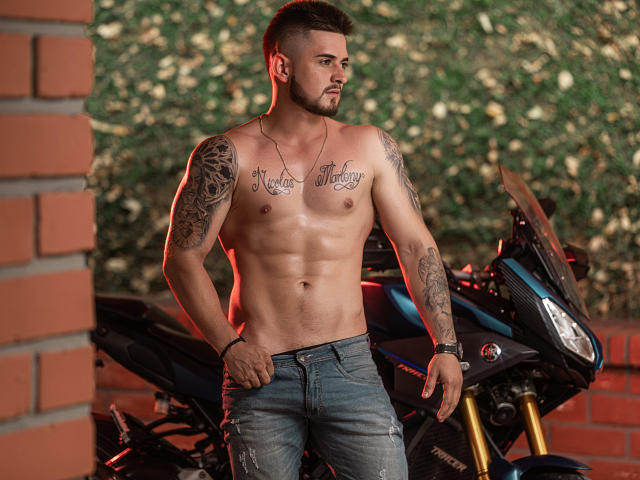 Ashton MULLER sem camisa, com tatuagens no peito e nos braços, calça jeans azul, em pé ao lado de uma motocicleta, olhando para o lado, cabelo castanho curto, barba, pulseira no pulso direito..