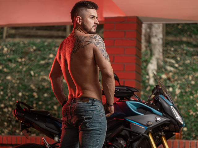 AshtonMuller ohne Hemd, mit Tattoos, in blauen Jeans, stehend mit den Händen in den Hüften, zurückblickend, neben einem schwarzen Motorrad, rotes Licht, kurze braune Haare, muskulös, ernster Gesichtsausdruck..