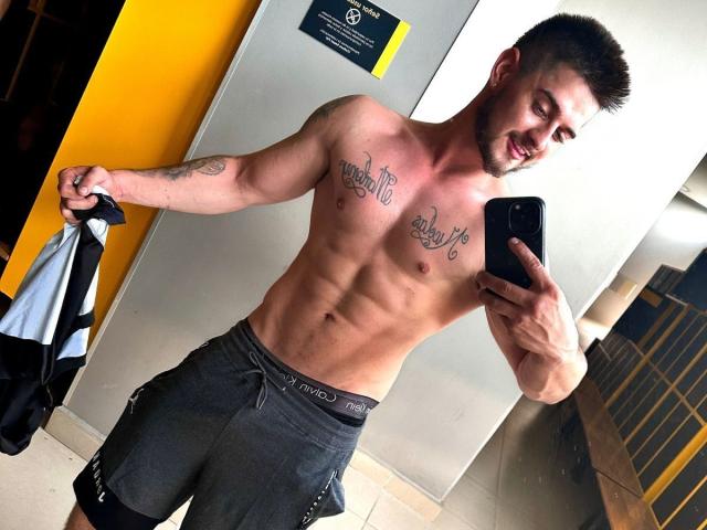 AshtonMuller, sem camisa, musculoso, com tatuagens no peito, segurando um celular, shorts cinzas, segurando um casaco, cabelo curto e escuro, sorrindo, selfie no espelho, abdômen visível, pele bronzeada, em um ambiente que se assemelha a uma academia..