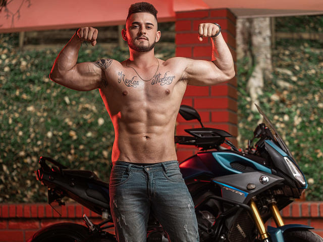 AshtonMuller, ohne Hemd, muskulös, die Muskeln anspannend, Tätowierungen auf der Brust, blaue Jeans, steht vor einem blauen Motorrad, Hintergrund in Rot und Backstein, kurze, dunkle Haare, Bart, ernster Gesichtsausdruck..