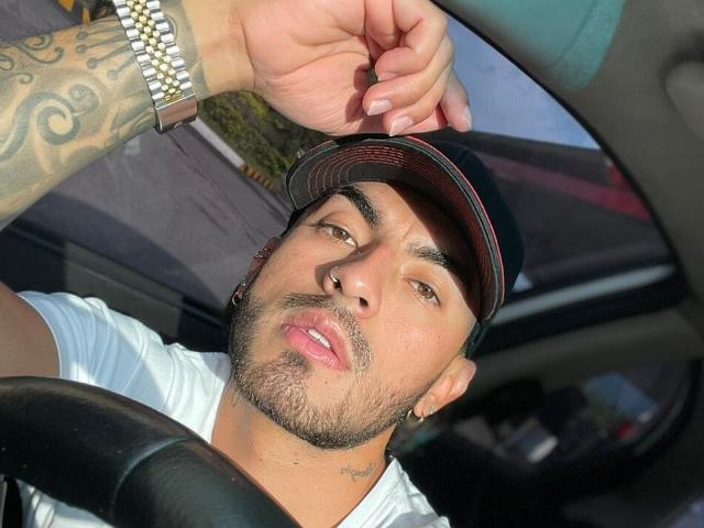 Zhion, with tattoos on his arm, wearing a baseball cap and white shirt, has his hand on his head, sitting in a car, looking at the camera with a slightly open mouth.