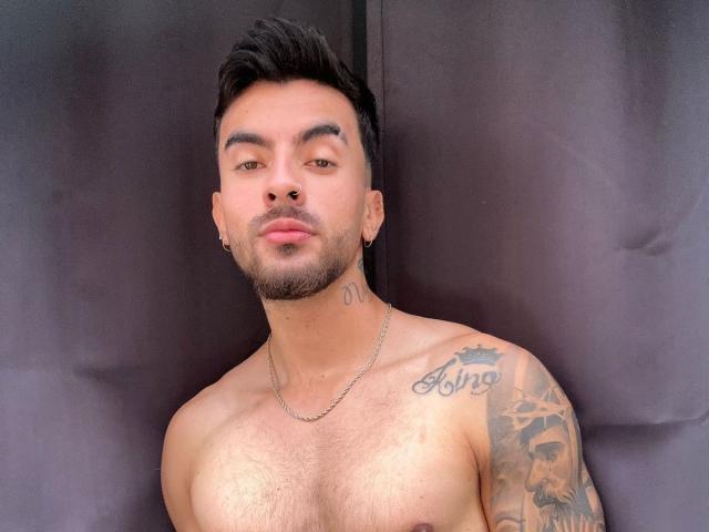 Zhion, met kort zwart haar, lichtbruine huid en een baard, zonder shirt, toont een tatoeage op zijn rechter schouder en een ketting om zijn nek. Zijn borst is behaard en hij kijkt naar de camera..